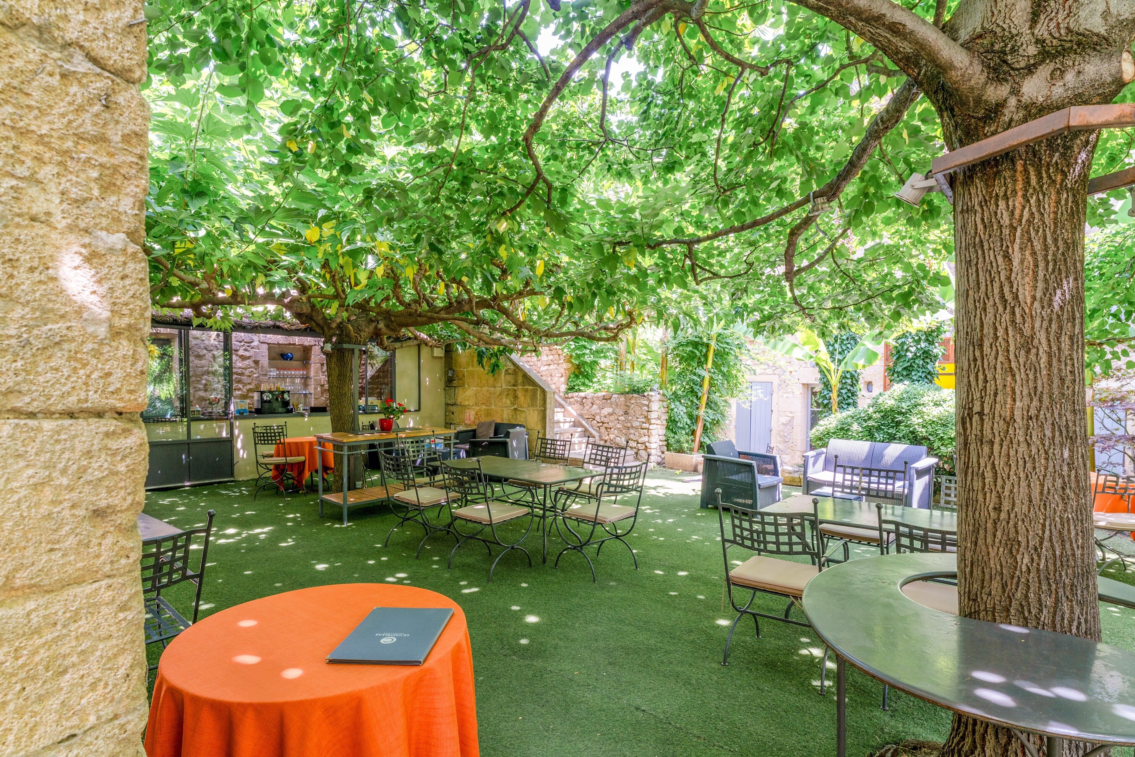 An outdoor patio area with tables and chairs, surrounded by trees and a stone wall.