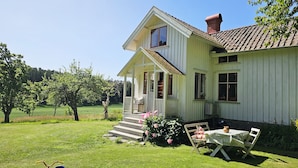 A white house with a porch, a small table with a chair, and a garden with trees and grass. - Charming house with kayaks, 300m from the sea on Orust | SE09139 (Henån)