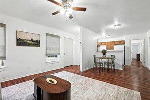 A spacious living room with hardwood flooring, a ceiling fan, a dining area with a table and chairs, and a kitchen in the background.