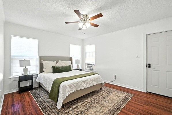 A bedroom with a bed, a ceiling fan, a window, a door, and a nightstand with a lamp.