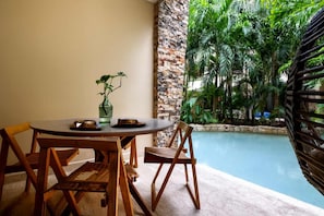 A dining area with a round table and wooden chairs, a potted plant, and a view of a swimming pool surrounded by lush greenery.