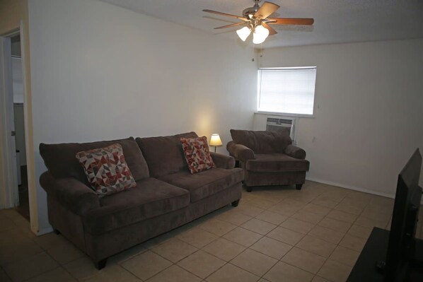 A living room with a brown sofa, a smaller armchair, a ceiling fan, and a television.