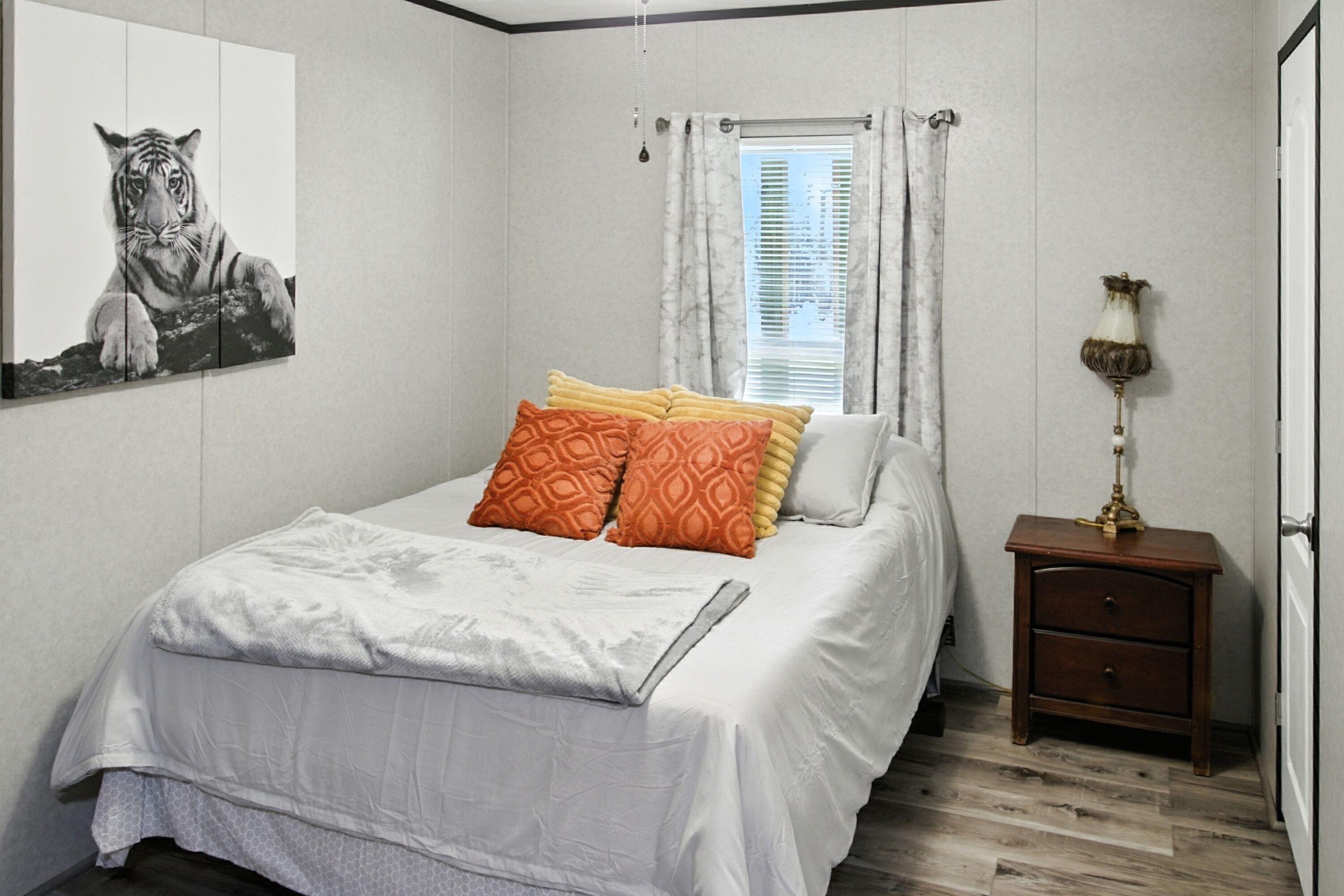 A bedroom with a bed, pillows, a nightstand, a lamp, a window with curtains, and a tiger painting on the wall.
