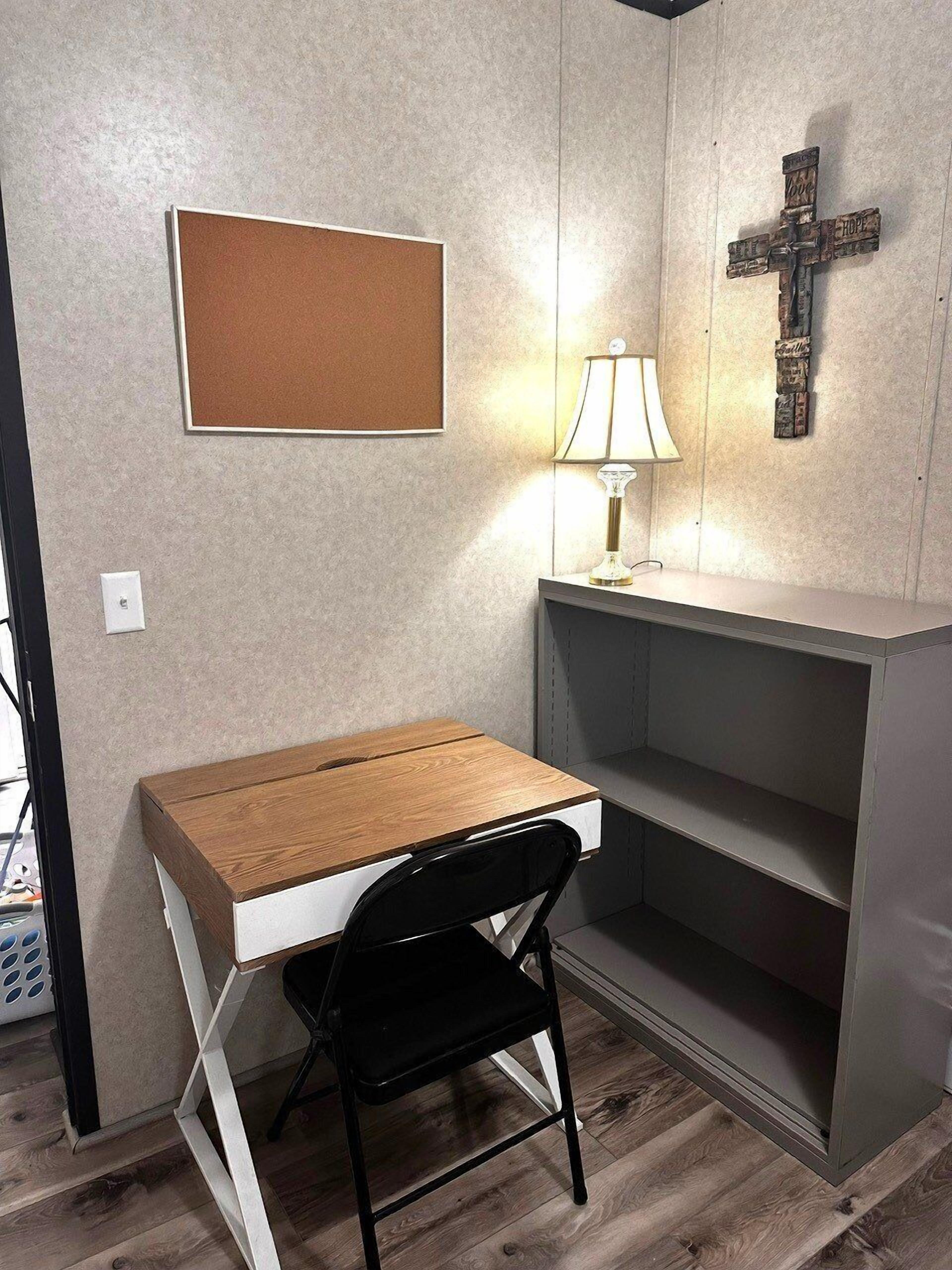 A small, private office space with a wooden desk, a black chair, a corkboard, a lamp, and a religious cross on the wall.