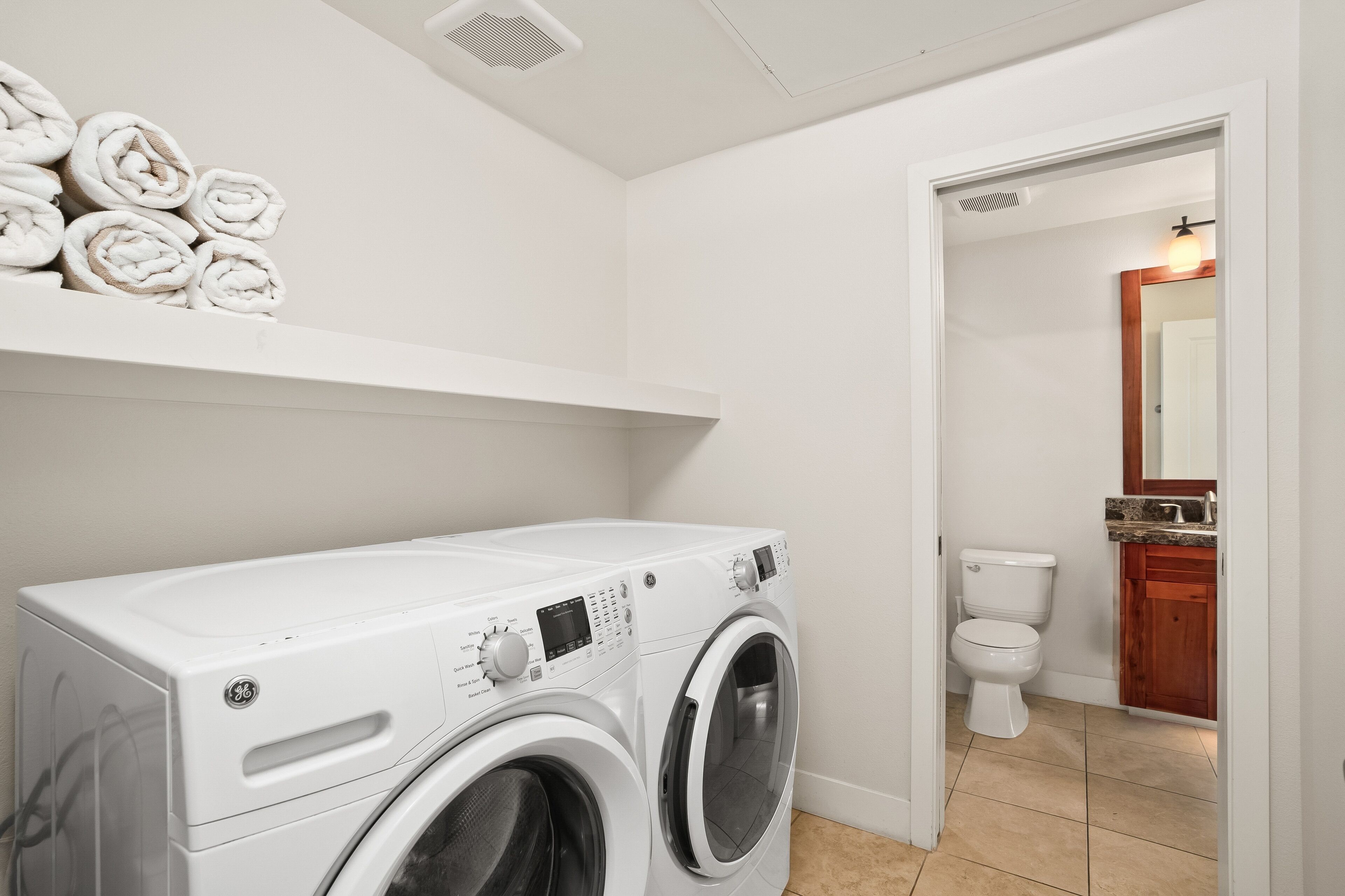 A laundry area with a front-loading washer and dryer, a shelf with rolled towels, and a view into a bathroom with a toilet and sink.