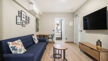 Una sala de estar moderna con un sofá, una mesa de centro y un televisor. Se ve un comedor con una mesa y sillas, y una cocina visible a través de una puerta abierta.