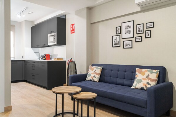 A modern living room with a blue sofa, two wooden side tables, and a kitchen area with grey cabinets and a microwave. - Dobo Madrid Cabrera (Madrid)