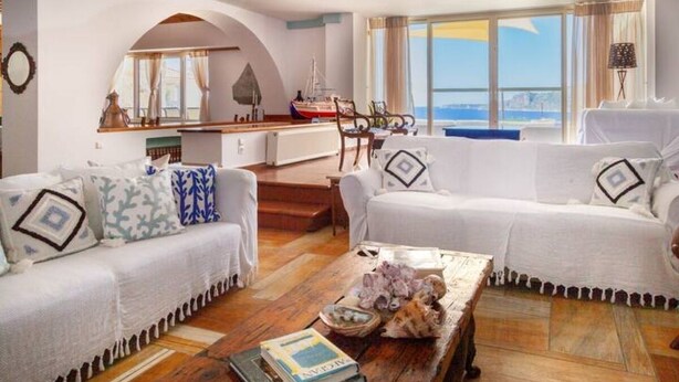 Una sala de estar moderna con dos sofás blancos, una mesa de centro de madera y vista al mar a través de grandes ventanales.