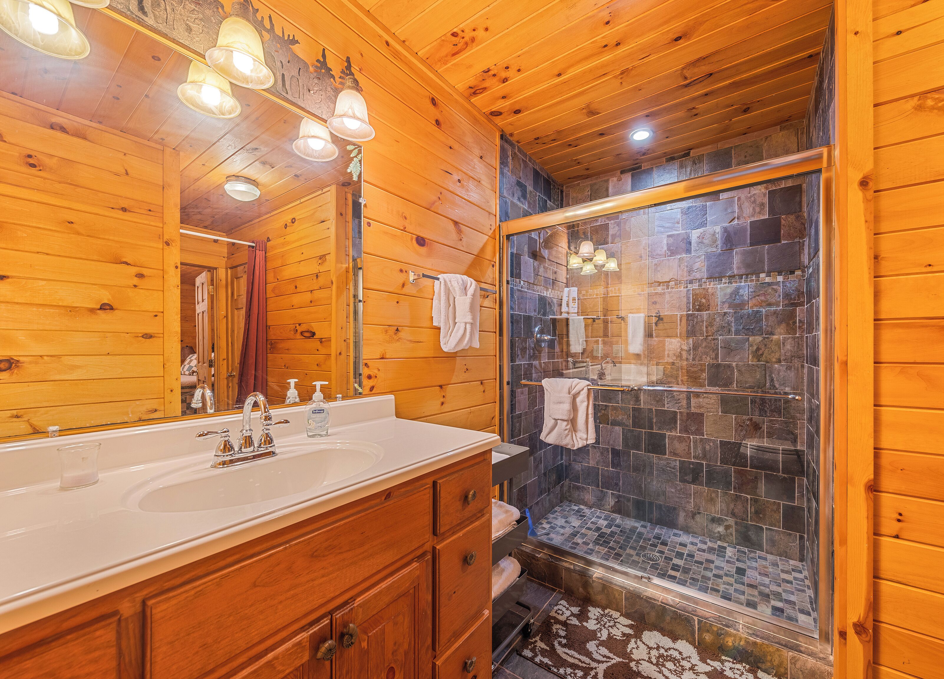 A wooden cabin bathroom with a large mirror, a sink with two faucets, a glass shower enclosure, and a wooden vanity with drawers.