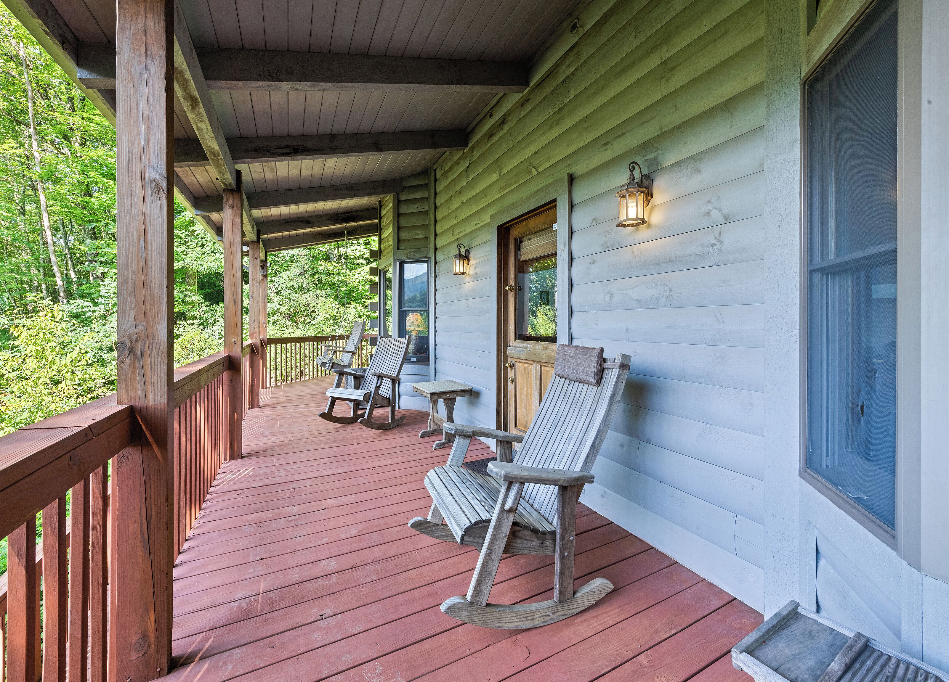 A wooden porch with rocking chairs and a table, overlooking a forested area.