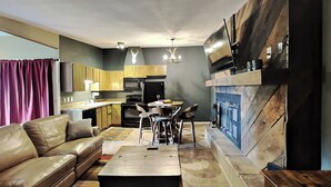 Una sala de estar moderna con sofá, mesa de centro y chimenea. Se ve la cocina al fondo, con armarios de madera y un comedor.