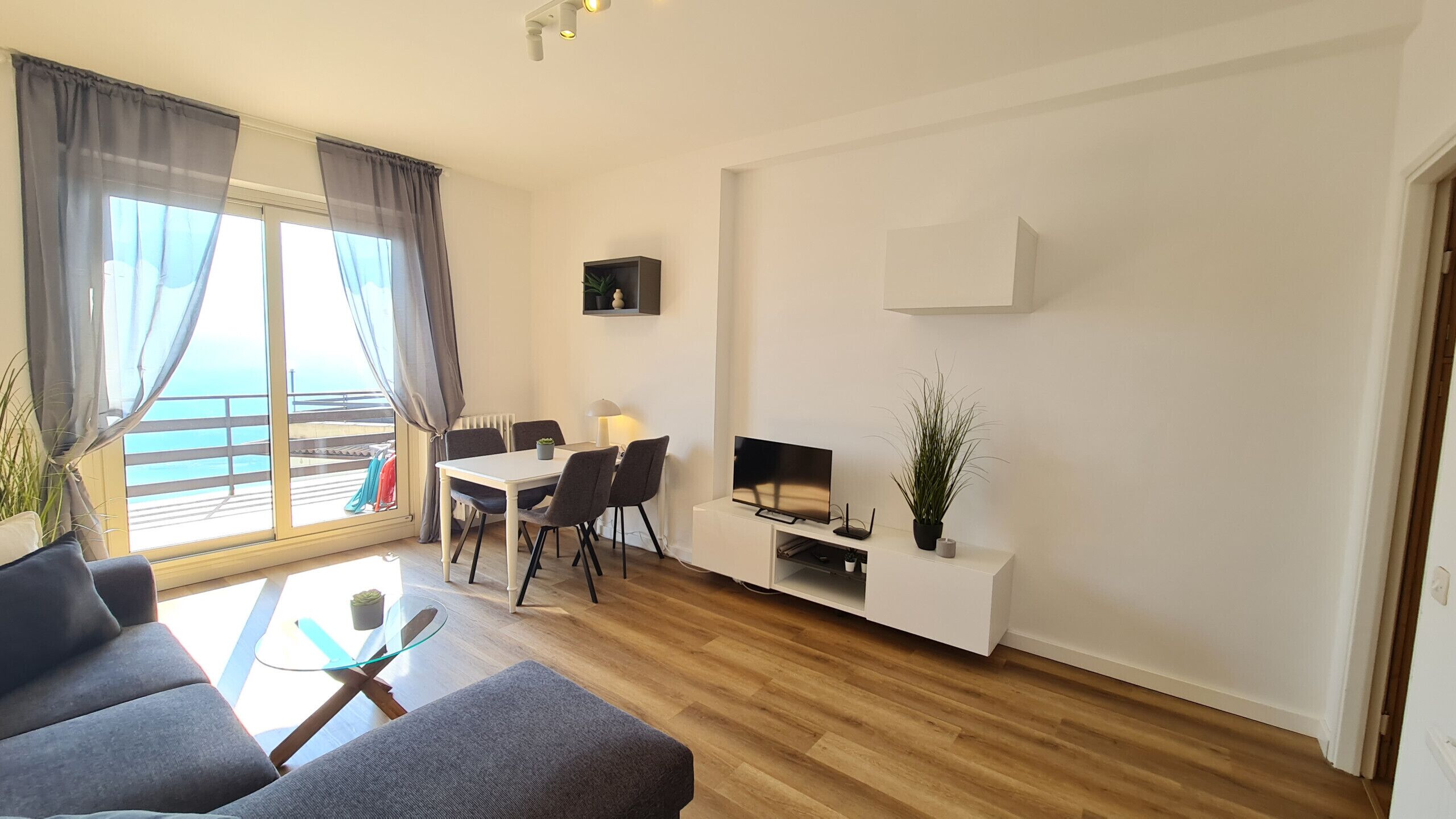 A modern living room with a sofa, a glass coffee table, a dining table with chairs, a flat-screen TV, and a potted plant.