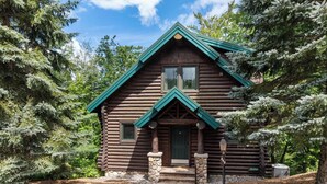 A log cabin with a green roof, stone foundation, and a small porch with steps leading to the door.