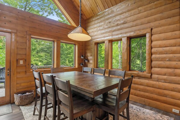 A wooden cabin interior with a dining table and chairs, large windows, and a hanging light fixture.