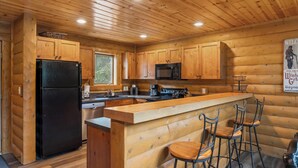 A wooden cabin-style kitchen with a black refrigerator, a microwave, a coffee maker, and a bar area with stools.
