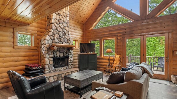 A log cabin interior with a stone fireplace, wooden furniture, and large windows.