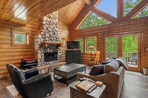 A log cabin interior with a stone fireplace, wooden furniture, and large windows.