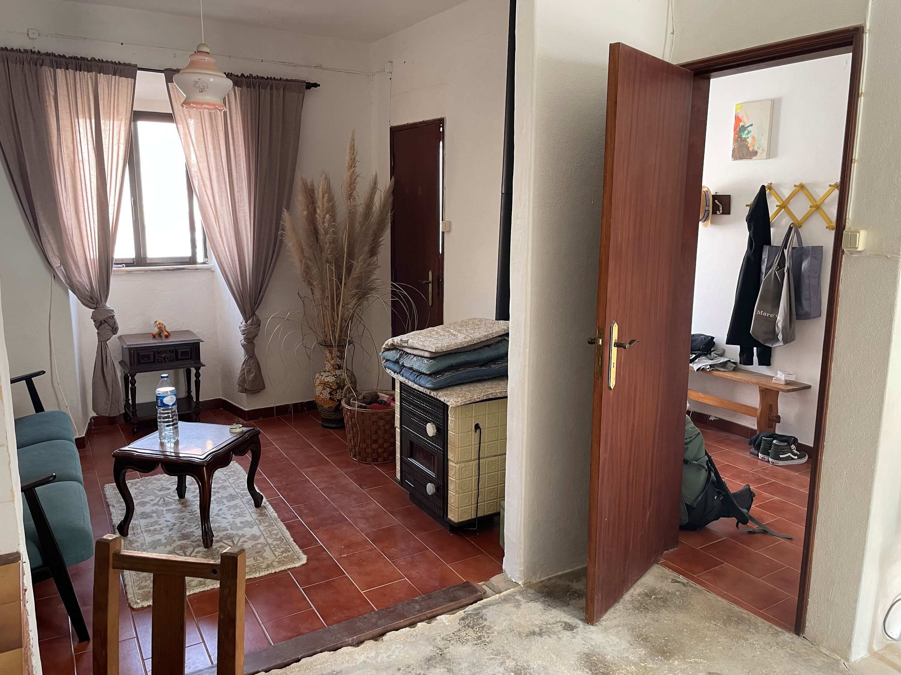 Une pièce comprenant un canapé, une petite table, un vase décoratif, une bouteille d’eau et une porte donnant sur une autre pièce où se trouvent un banc et un porte-manteau.