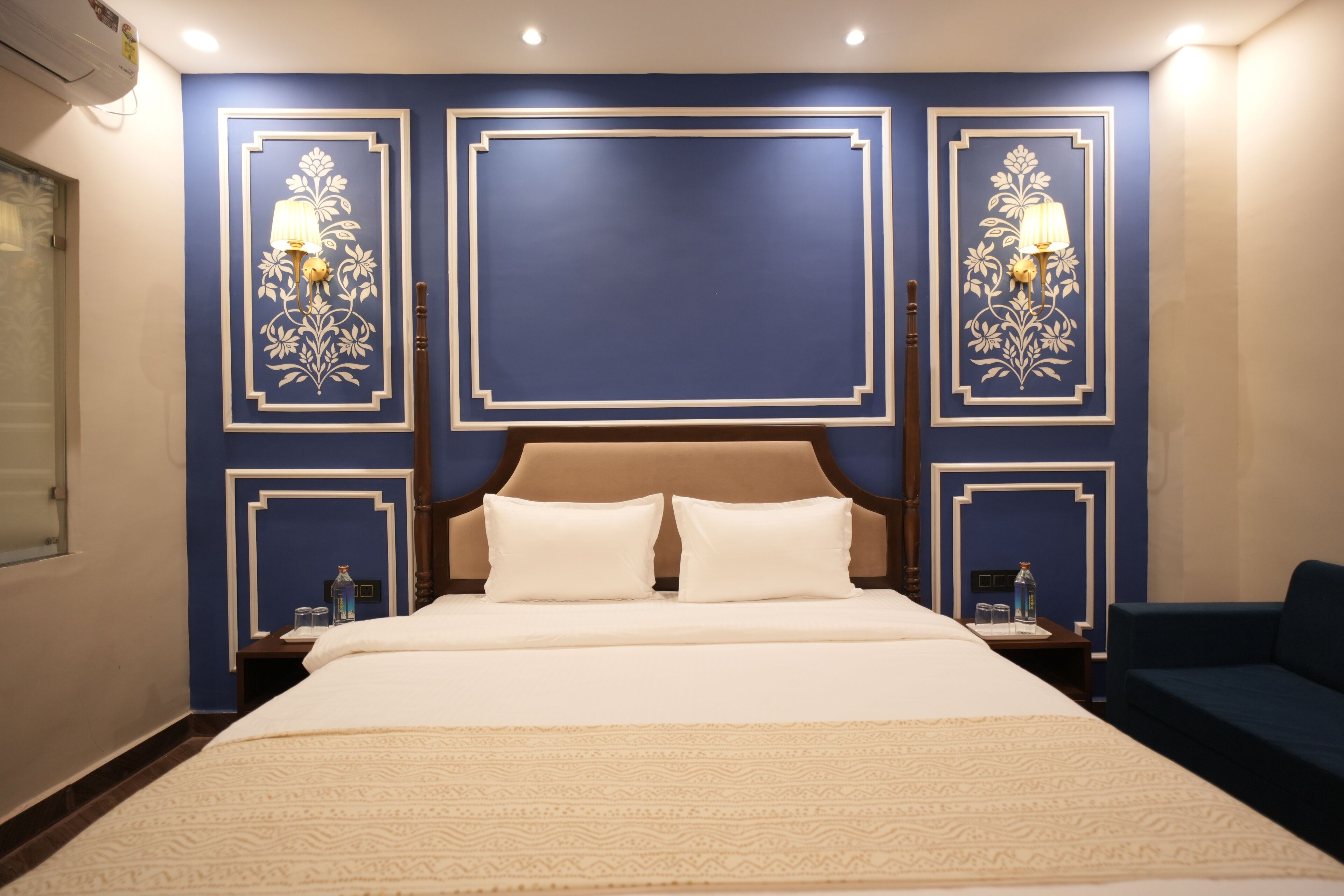A hotel room with a large bed, two bedside tables, a nightstand, a blue wall with decorative panels, and a blue sofa.