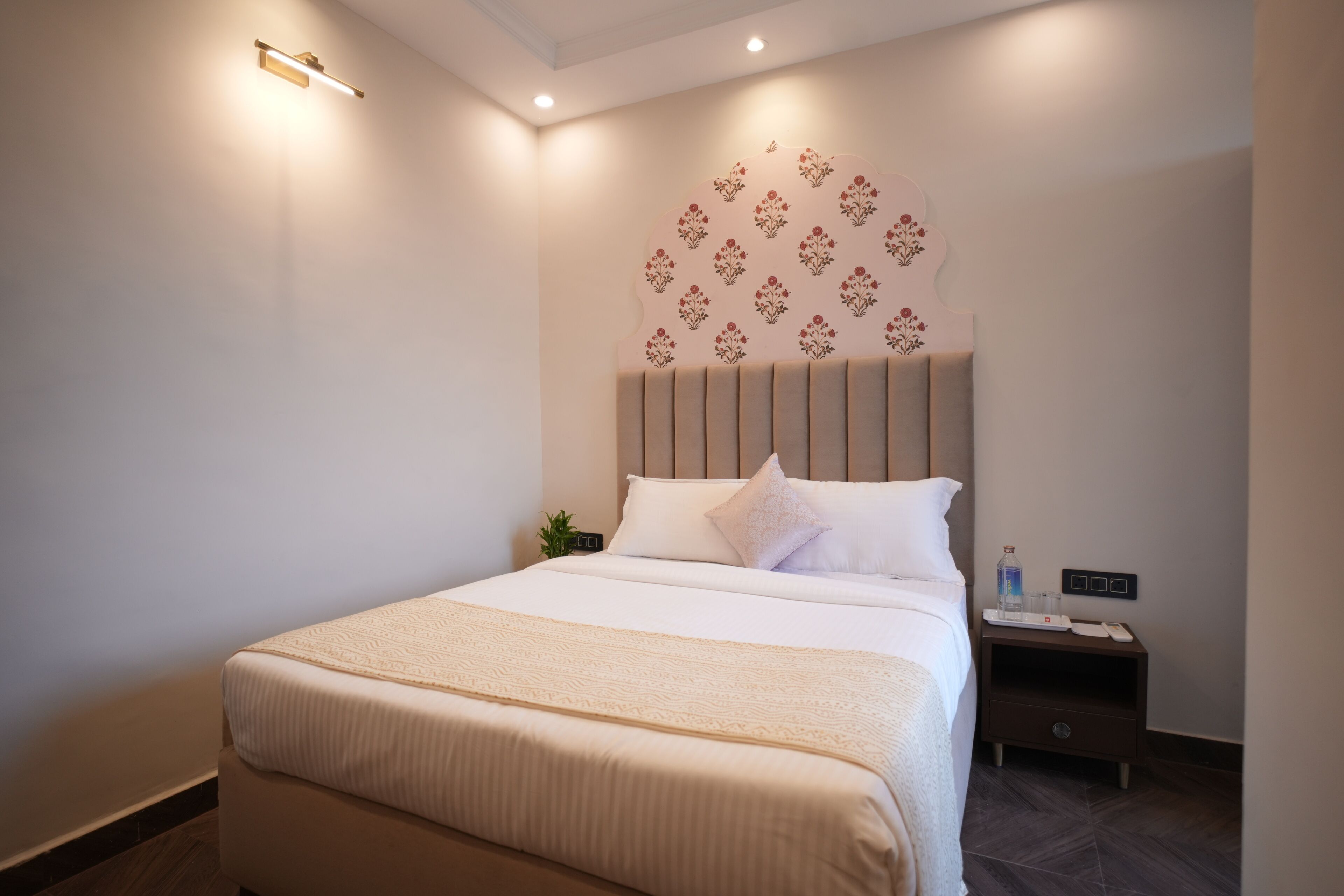 Una cama bien hecha con cabecera, una mesita de noche con lámpara, y un aplique en la pared.
