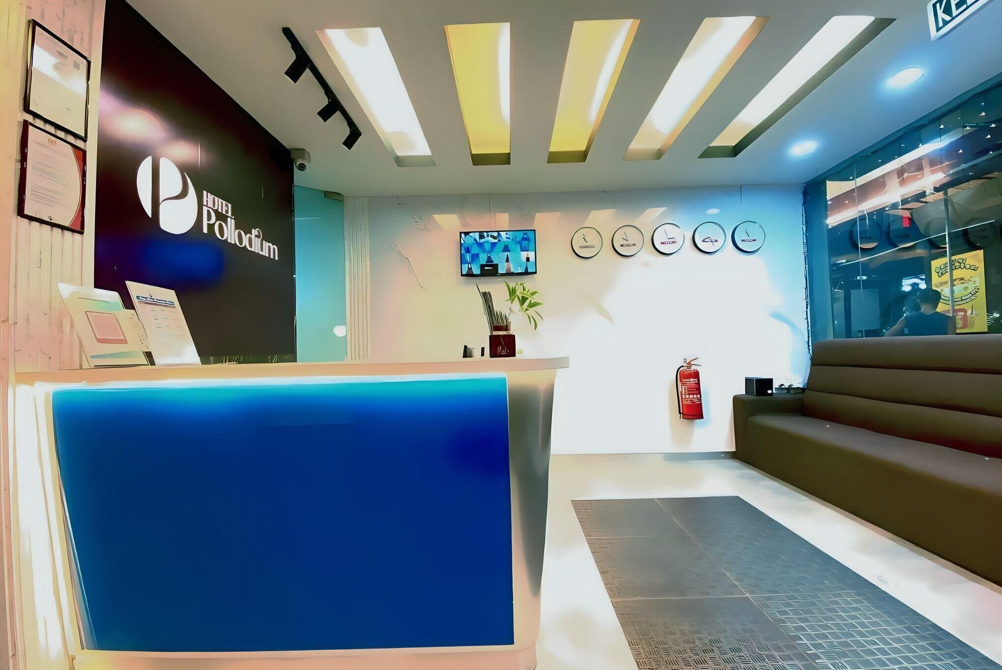 A hotel reception area with a blue reception desk, a black wall with the hotel name, a TV screen, and a fire extinguisher.