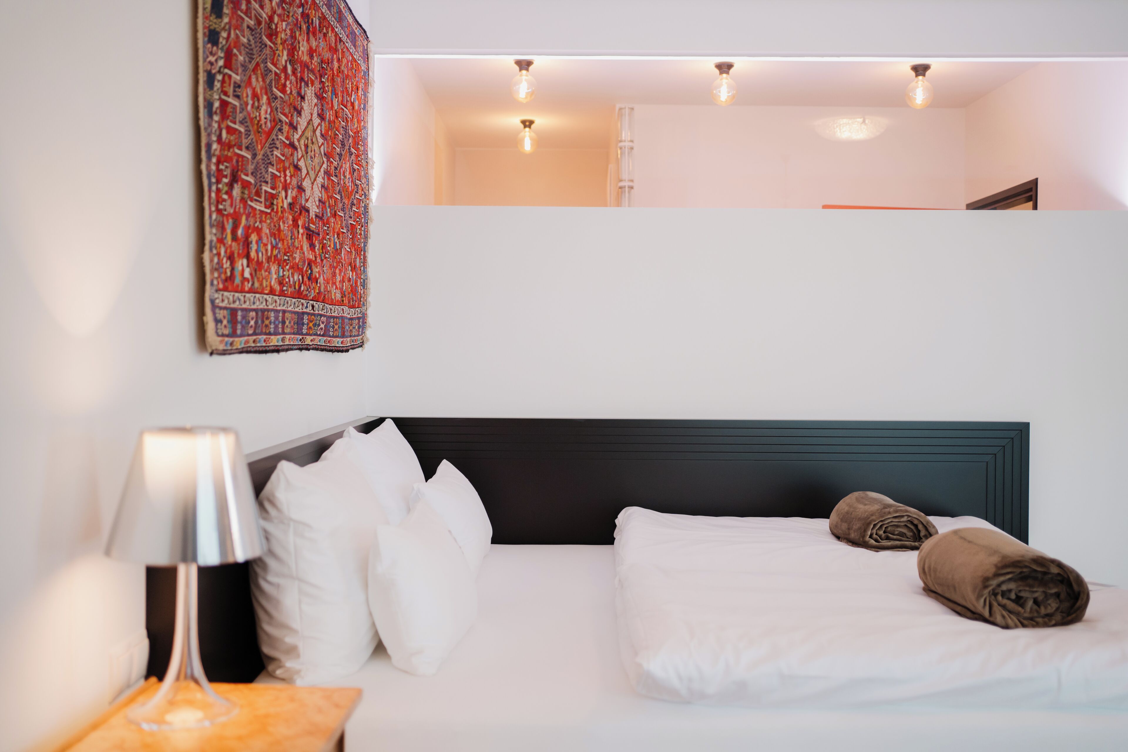 Een opgemaakt bed met witte en bruine lakens, een bedlamp en een wandtapijt.