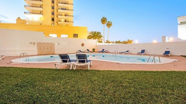 A swimming pool with lounge chairs, a tall building in the background, and palm trees.