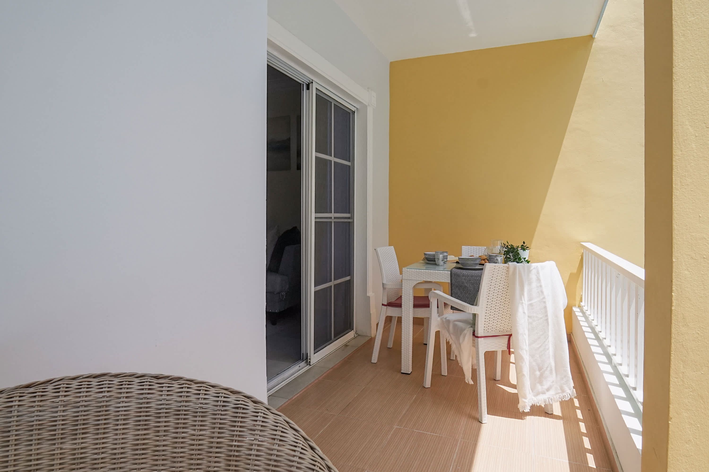 Een balkon met een tafel en stoelen, een rieten stoel en een schuifdeur met glas.