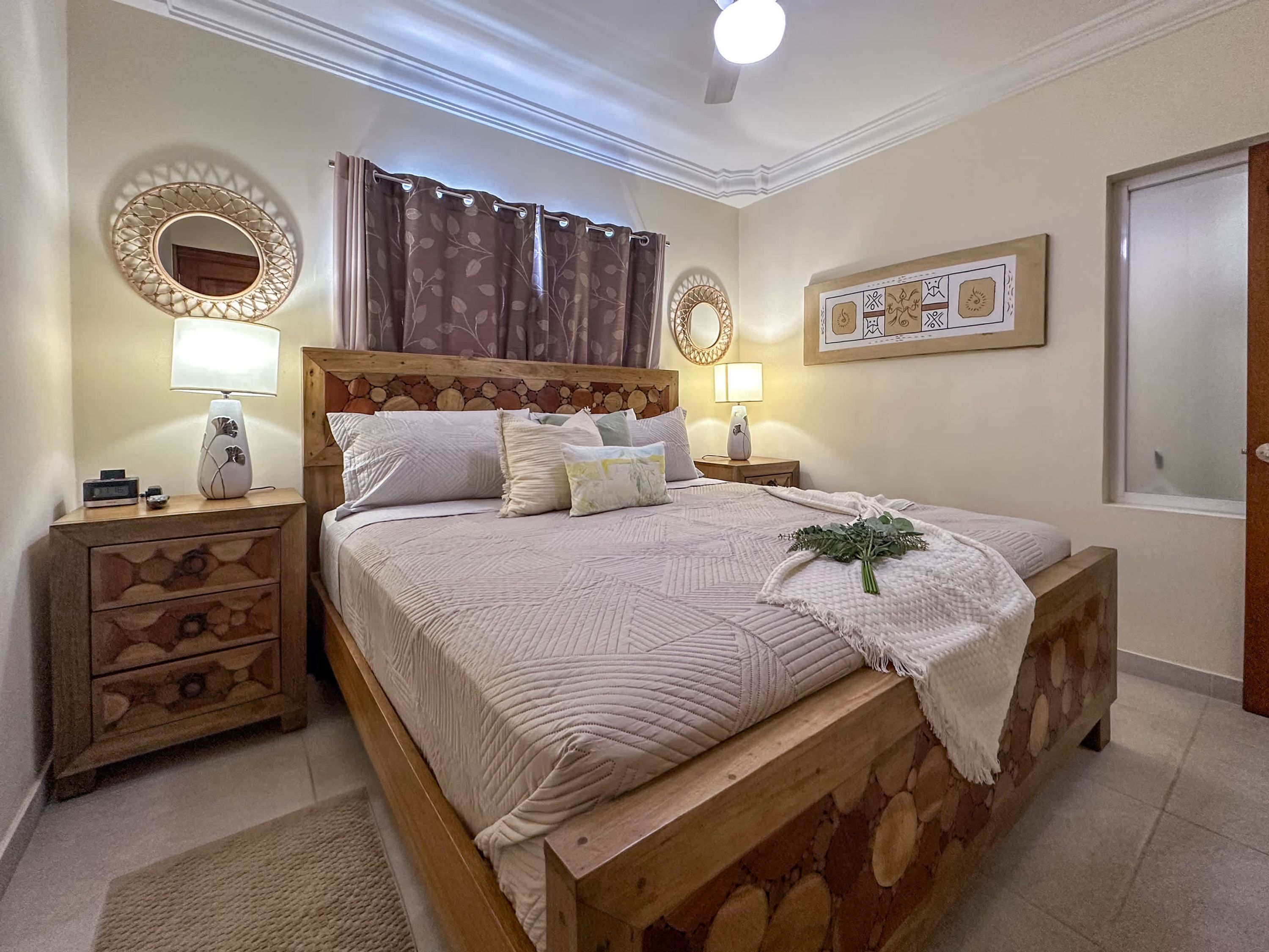 Un dormitorio con cama de madera, dos mesitas de noche con lámparas, una mesita auxiliar y un espejo decorativo, además de un cuadro en la pared.