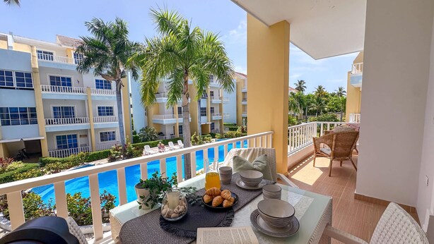 A balcony with a view of a swimming pool, lounge chairs, dining table with plates and a book, and a potted plant.