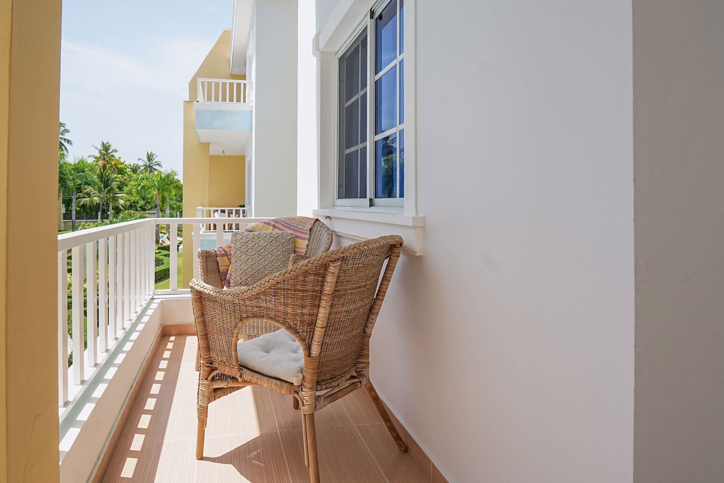 A balcony with a wicker chair, a white cushion, and a window overlooking a garden.