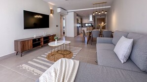 A modern living room with a grey sofa, a flat-screen TV, a wooden entertainment unit, and a dining area with a kitchen in the background. - Beautiful Brand new Beach Condo w Picuzzi (Punta Cana)
