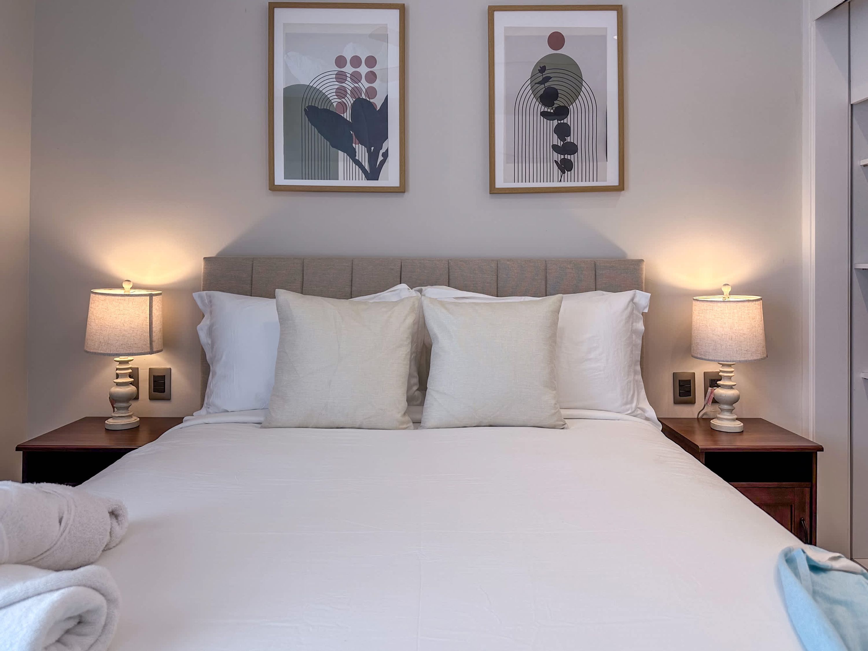 A neatly made bed with white linens, two bedside lamps, and two framed artworks on the wall.