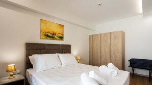Una cama bien hecha con sábanas blancas y toallas enrolladas, un armario de madera, un escritorio con lámpara y un cuadro en la pared.