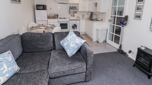 En kompakt opholdsstue med en grå sofa, et spisebord med stole, et køkken med hvide skabe, en vaskemaskine og en lille, sort komfur.