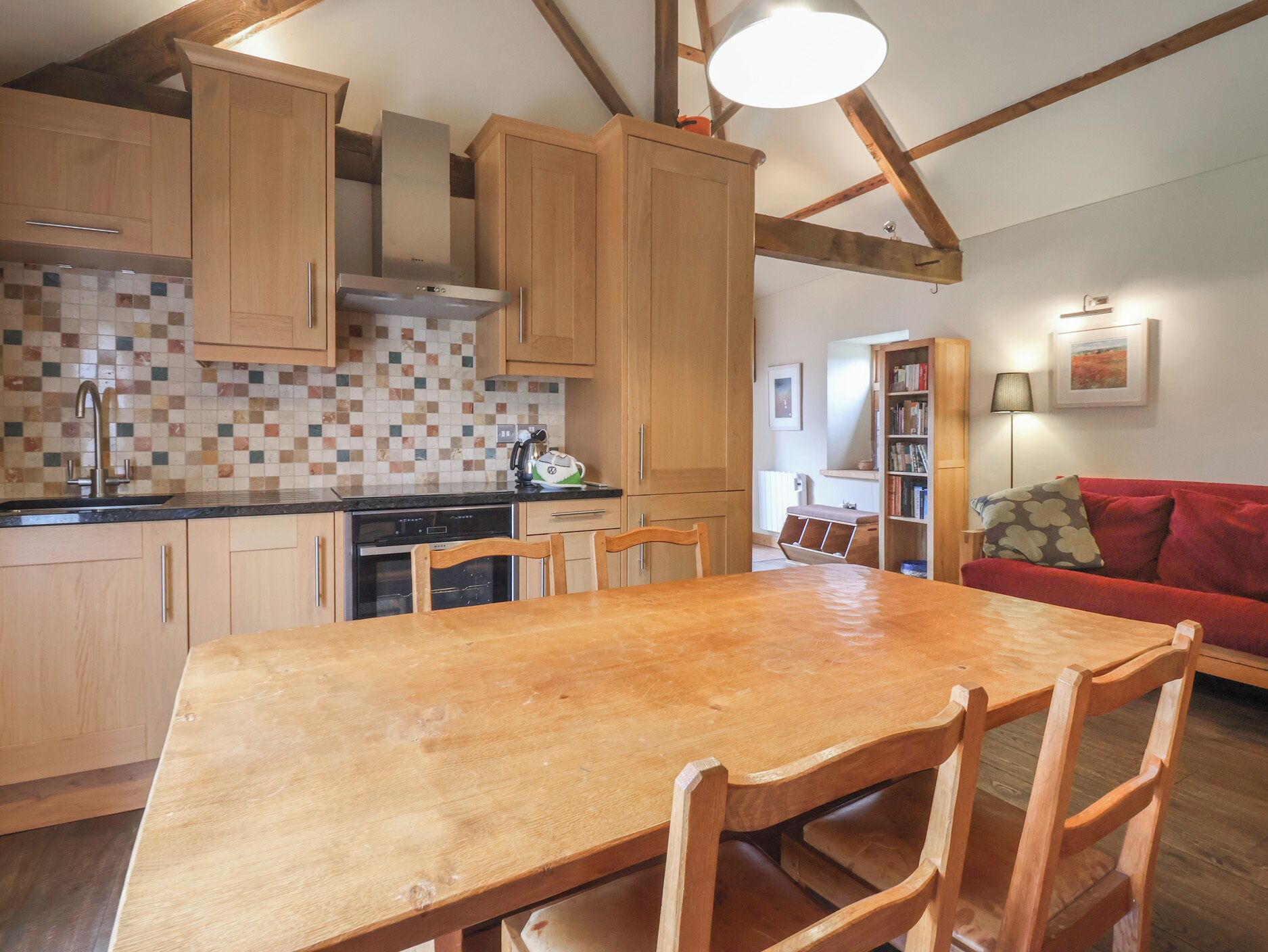 A kitchen with wooden cabinets, a tiled backsplash, a dining table with chairs, and a red sofa in the living area.