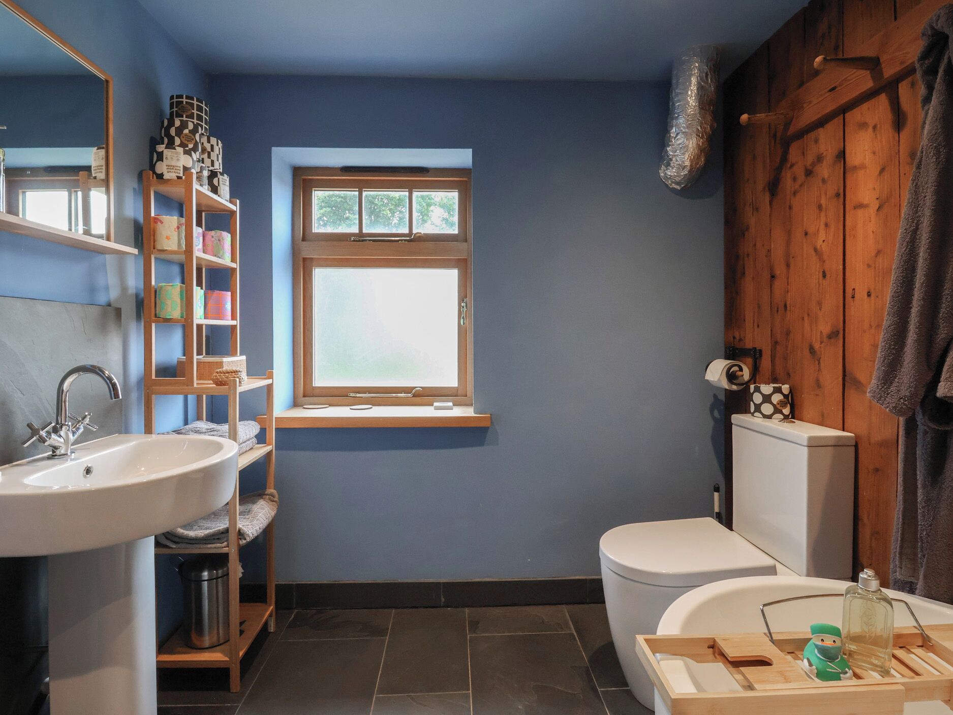 A bathroom with a white sink, a toilet, a window with a view, and a shelf with toiletries.