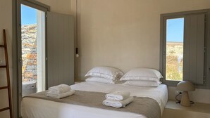 A bedroom with a bed, two windows, a lamp, and a view of the outdoors. - Pentanemi Folegandros (Folegandros)
