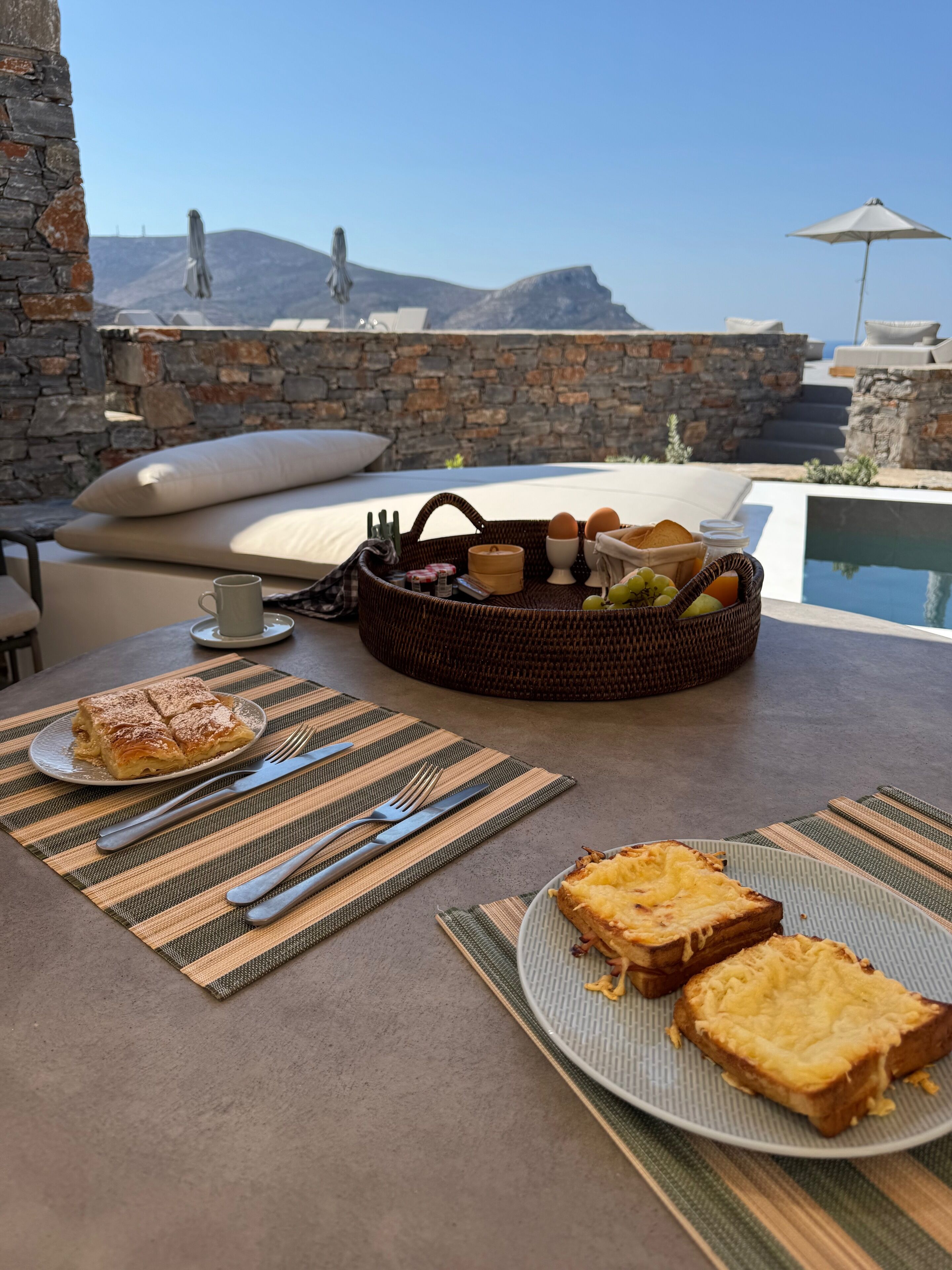 Esmorzar amb una cistella de pa, ous i fruita, un plat de torrades i una tassa en una terrassa amb piscina i parets de pedra al fons.