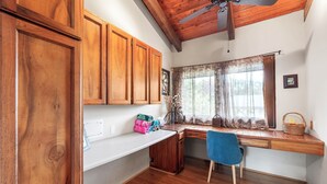 A kitchen with wooden cabinets, a built-in desk, a blue chair, and a window with curtains. - Architectural Gem With Infinity Pool Ocean Views - Vrkcv (Kailua-Kona)