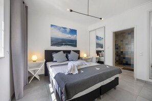 A modern bedroom with a large bed, bedside lamps, a view of the sea, and a tiled shower area.