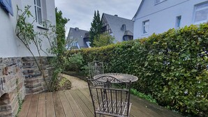 A wooden deck with a metal table and chair, surrounded by a hedge and a stone wall.