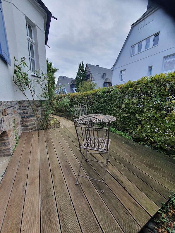 A wooden deck with a metal table and chair, surrounded by a hedge and a stone wall.