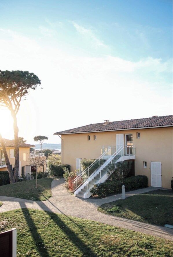 A residential building with a staircase leading to a balcony, surrounded by a well-maintained lawn and trees.