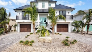 Una casa a due piani con un ingresso principale, due porte per garage e un giardino anteriore curato con palme e un elemento decorativo in pietra.