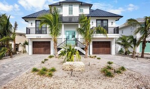 A two-story house with a front entrance, two garage doors, and a landscaped front yard with palm trees and a stone feature. - Beautiful Beach house! Heated pool and spa! (Holmes Beach)