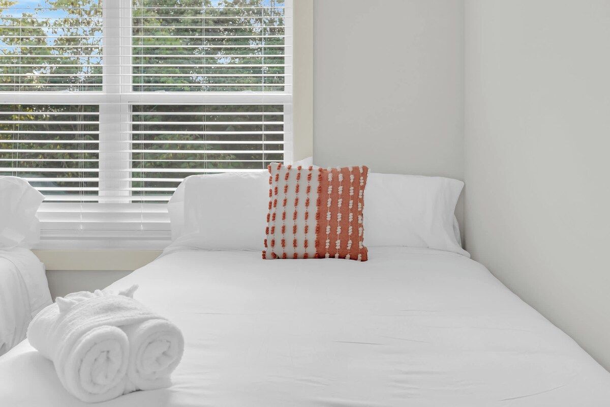 Una habitación de hotel con una cama, ropa de cama blanca, una ventana con persianas y una toalla blanca enrollada sobre la cama.