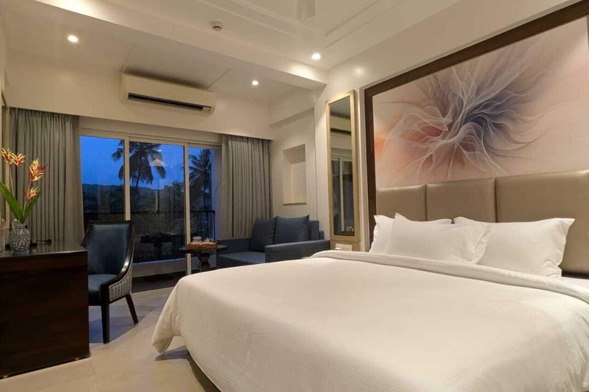 A hotel room with a large bed, a desk with a chair, a sofa, and a large window with a view of palm trees.