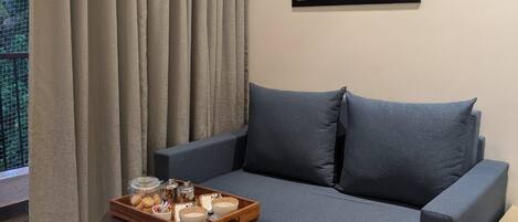 A modern living room with a grey sofa, a glass coffee table with a tray of snacks, and a framed abstract painting on the wall.