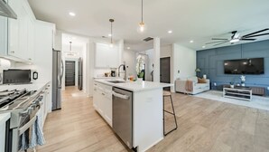 A modern kitchen with white cabinetry, a central island, and a dining area with a white sofa and a flat-screen TV. - 6 Beds| Center City | Bank of America Stadium (Charlotte)